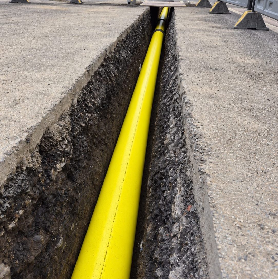 A gas main bedded in an open trench cut through a road surface, with pedestrian barriers set around the excavation